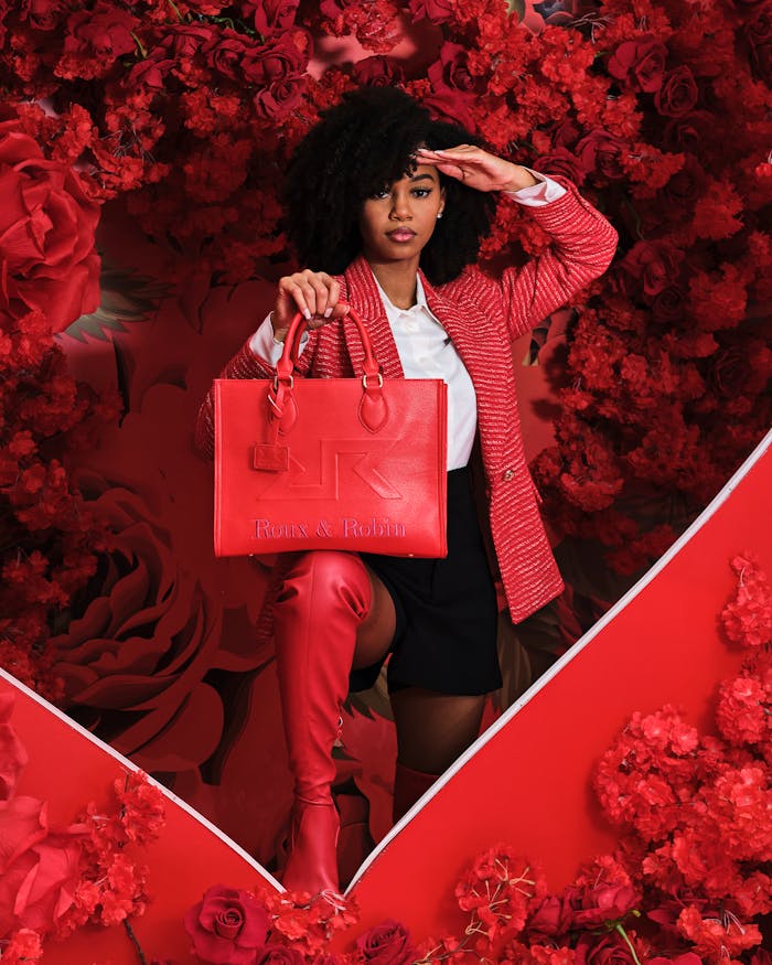 Stylish woman in red jacket and boots poses with red handbag, surrounded by roses, in a studio shoot.