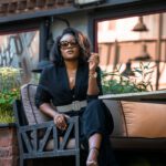 From below of trendy African American female in sunglasses sitting with crossed legs on outdoor couch in backyard and looking at camera