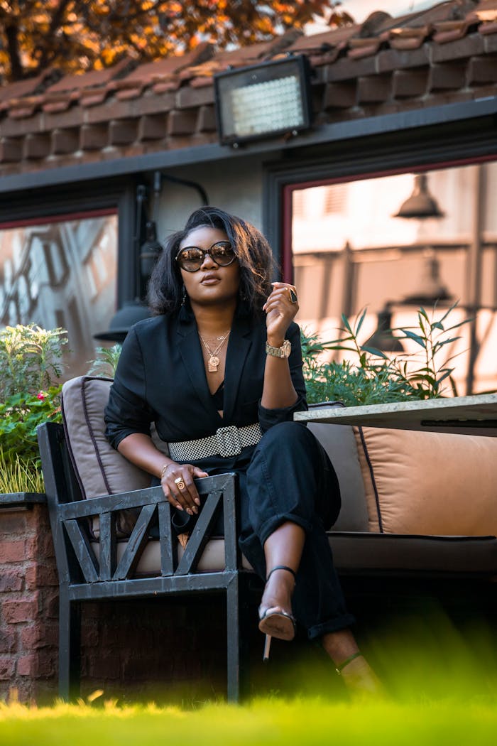 From below of trendy African American female in sunglasses sitting with crossed legs on outdoor couch in backyard and looking at camera