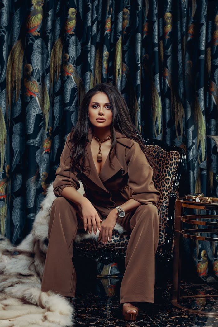Elegant woman in brown jumpsuit sitting on a leopard print chair in a richly decorated room.